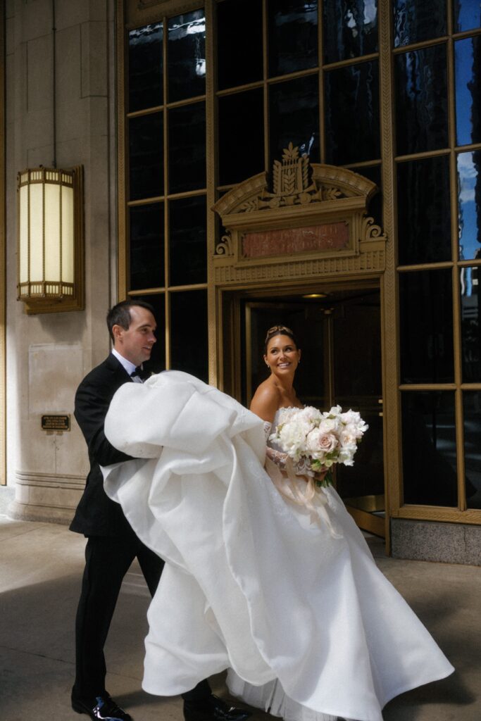 Bride and groom smile as groom holds her dress in downtown Chicago